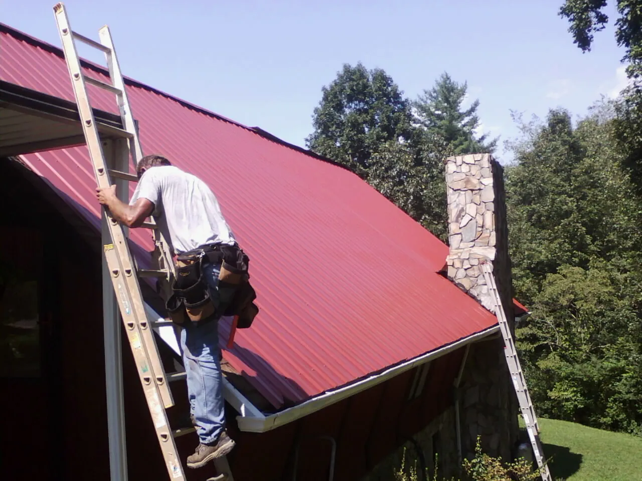 Roofer on a ladder beside a red metal roof and stone chimney — Soffit & Fascia Repair by Roofing Brothers in Oglesby